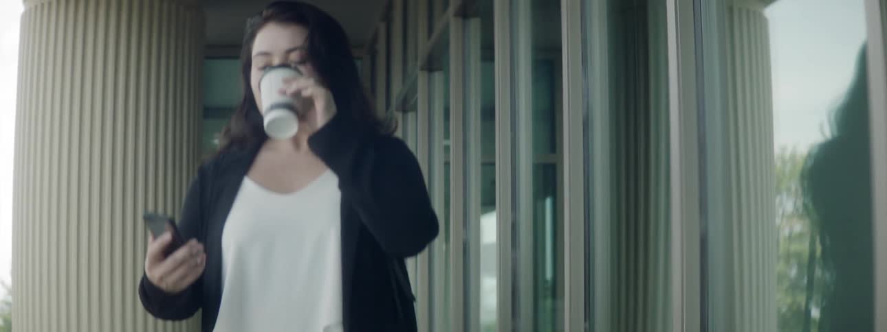 A woman walks on a sidewalk drinking a cup of coffee and checks her phone