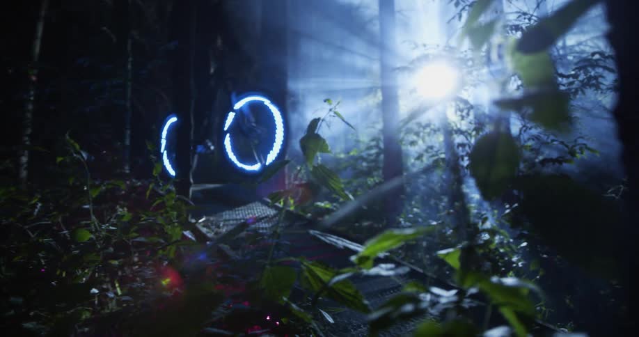 Person rides bicycle over narrow wooden pedestrian bridge under spotlight a dark forest