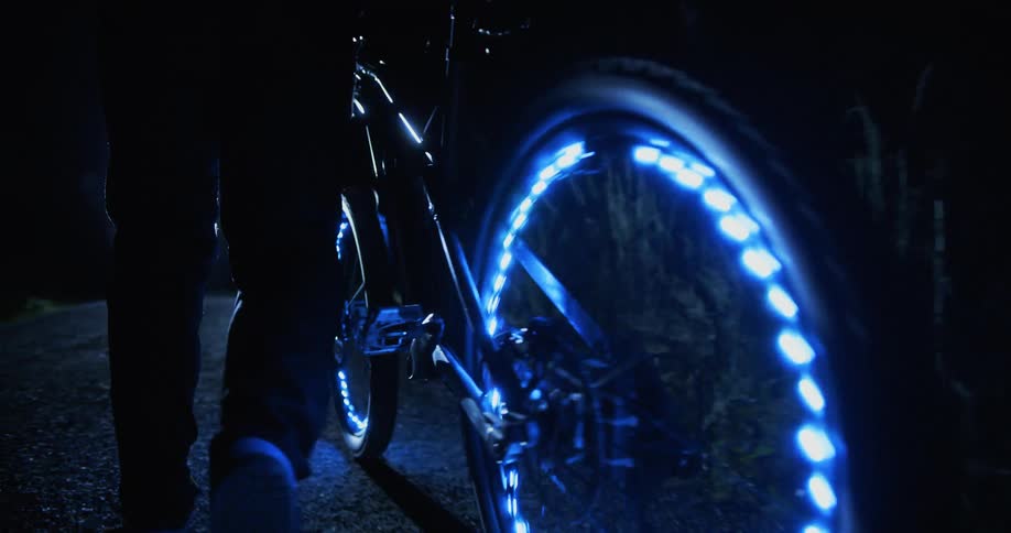 A person is pushing a bicycle at night that has illuminated wheels