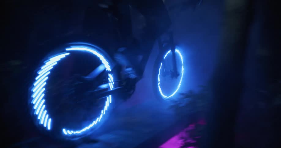A person rides a bike with blue led wheel lights through the trees and over a narrow bridge at night