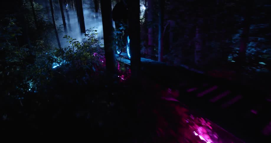A person riding a bicycle with glowing rims along a forest path outlined with neon lights