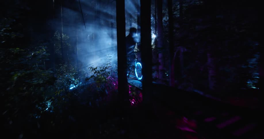 A spotlight shines through the trees at night as a person rides a bike with led wheel lights over a narrow bridge