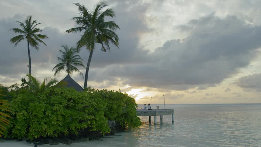 Two people sit on a small wooden dock over the ocean water as clouds pass overhead