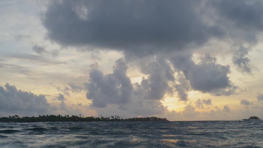 A dip beneath the rolling ocean waves with clouds in the sky and land on the horizon at sunset