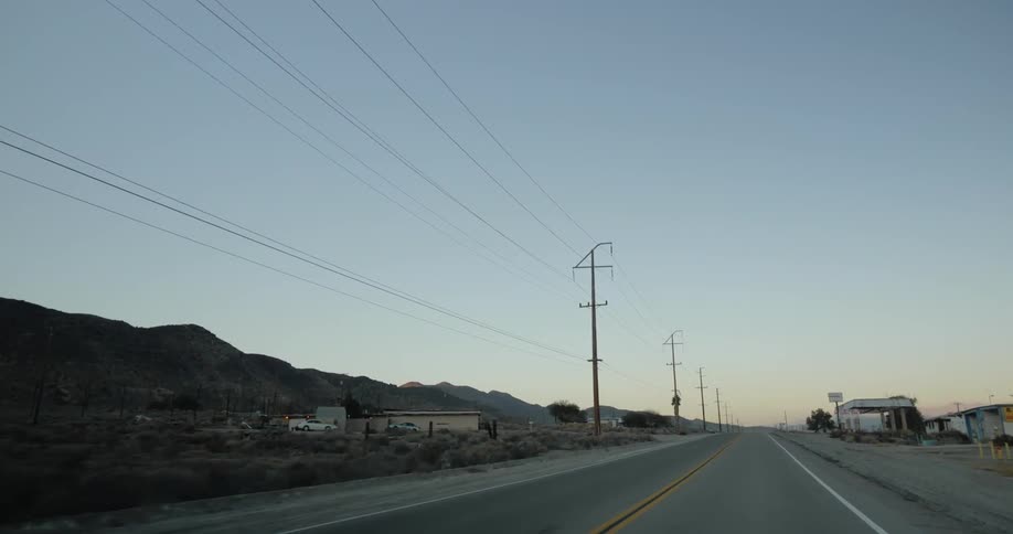 Traveling down road past buildings and scrub near hills