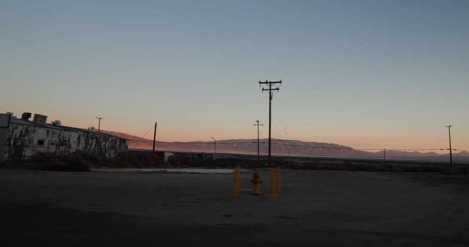 Driving past buildings in desert with mountains on the horizon as the sun sets