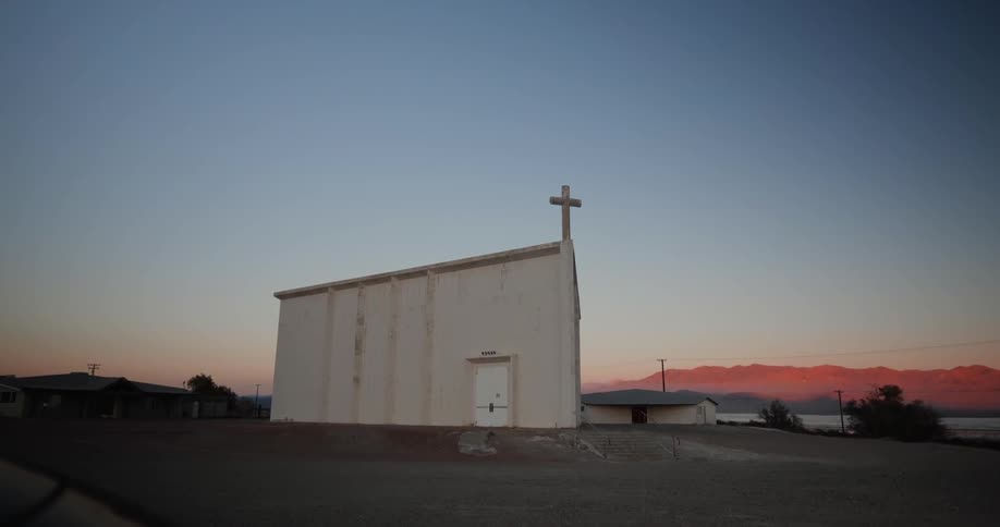 A cross stands on the corner of the rooftop of a plain white building in a rural area at sunrise
