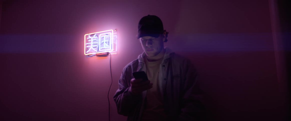 A man looking at his smartphone while standing next to a neon sign of chinese characters