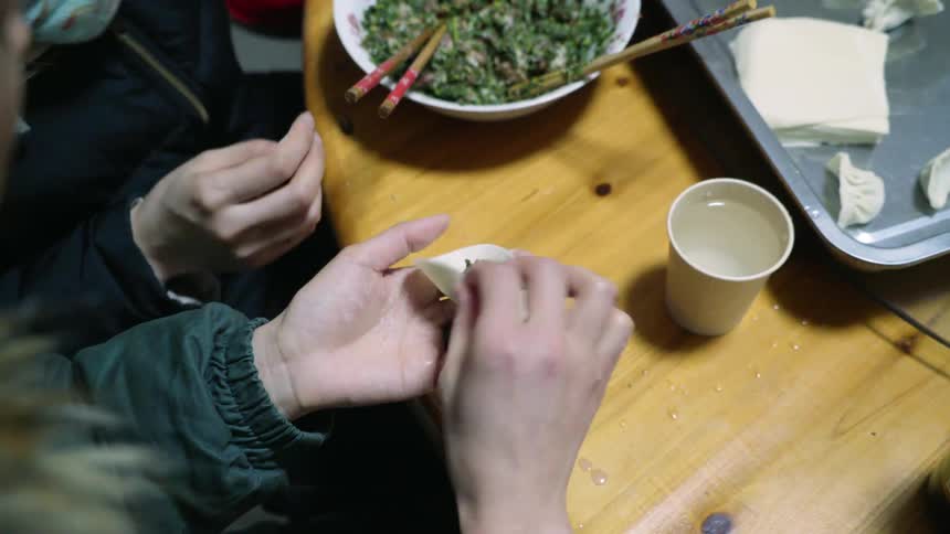 Women use chopsticks to add filling to round dumpling dough then seal the dumpling edges with fingers dipped in water