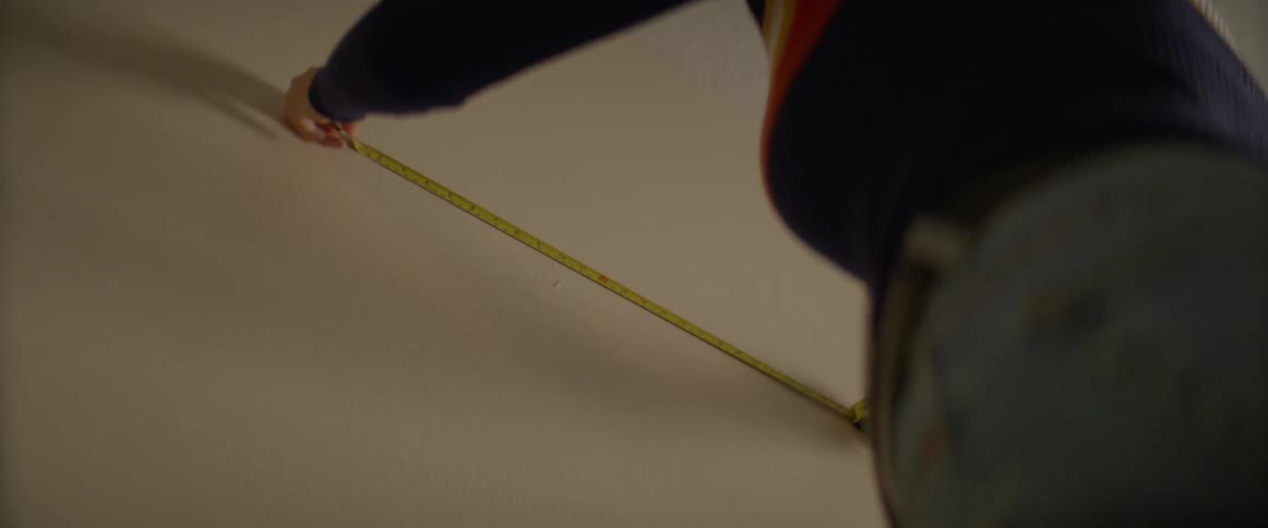 A woman stretches out a tape measure across an empty wall in order to measure the open space