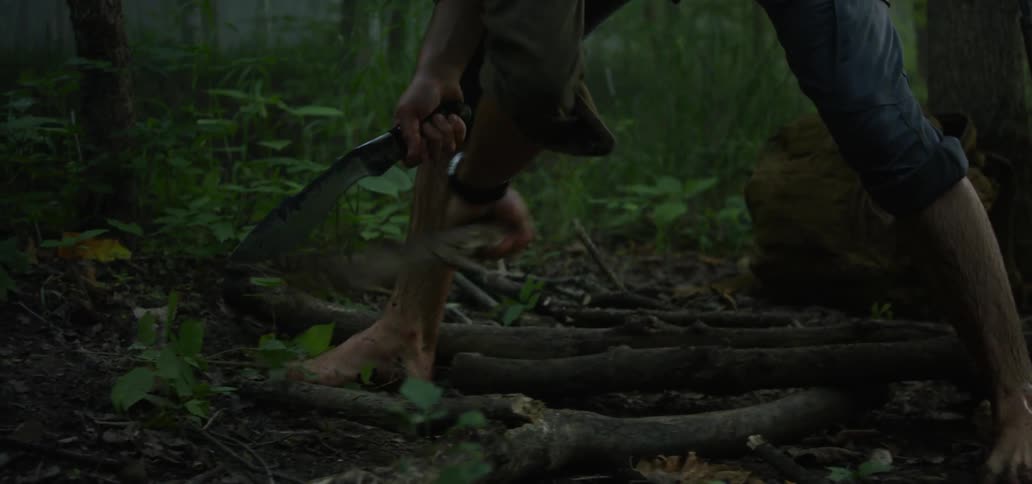 A man uses a machete to chop small logs in a wooded area