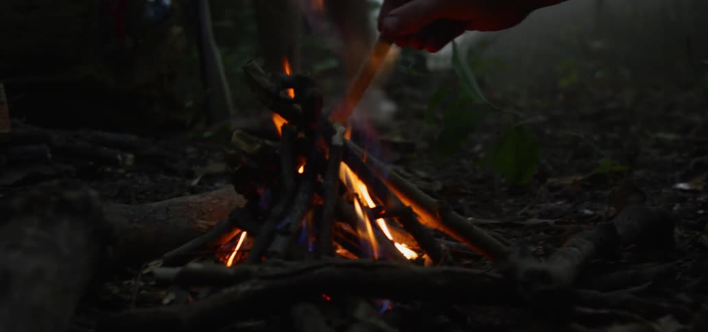 Two men in a forest work together to place sticks together in order to build up a camp fire