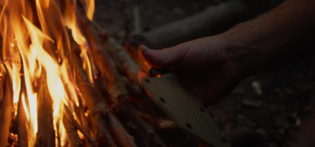 A man sits by a fire and examines a knife