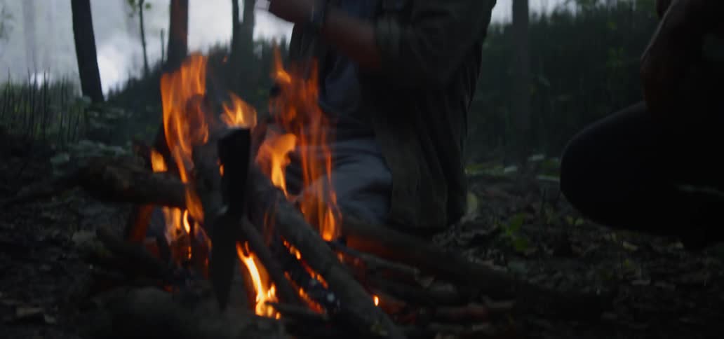 Two men are kneeling near a camp fire in the woods