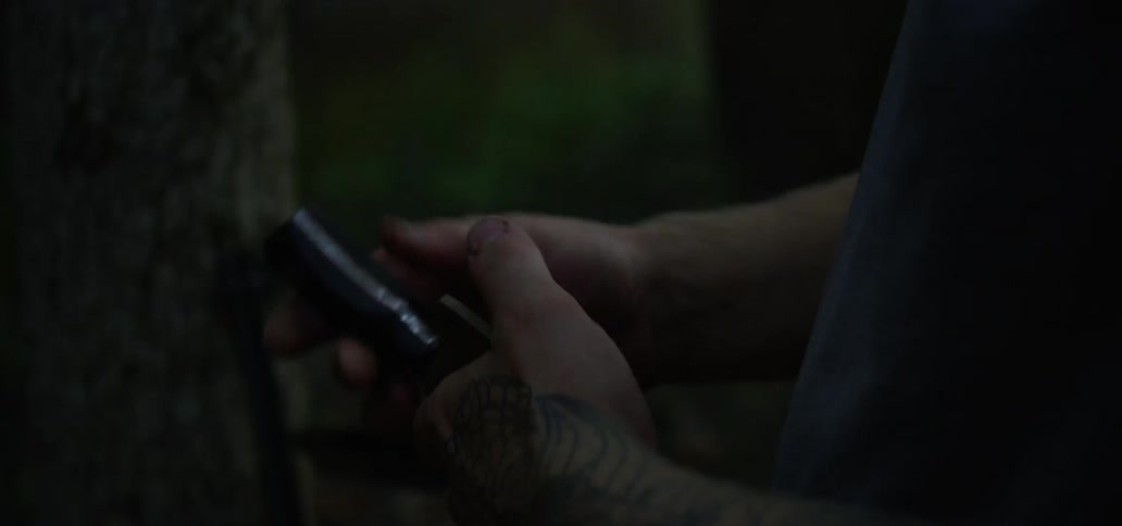 A man standing in a forest takes a knife out of its sheath and inspects it before shoving it back in