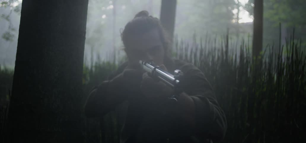 Man in crouched down in tall grass in the woods looking around through the scope of his rifle