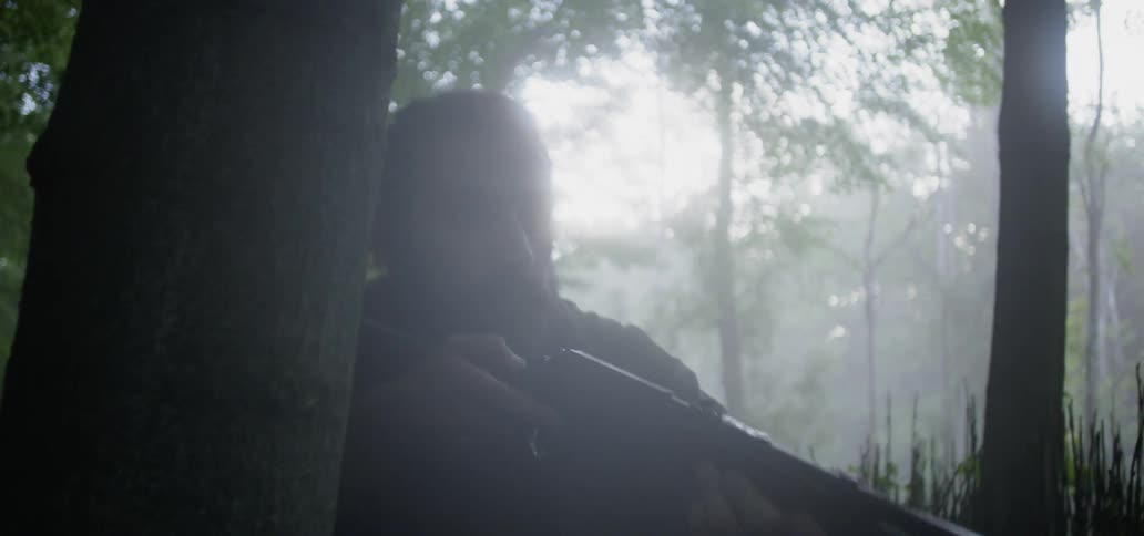 A man in a forest kneeling behind a tree to take aim with a rifle