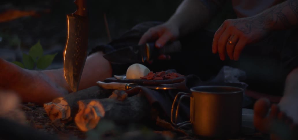A young male sits but a campfire in a forest while eating some food