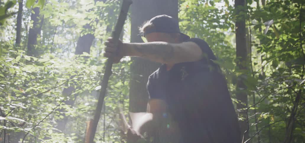 A man in a forest forcefully hacks away at a small tree in order to cut it down