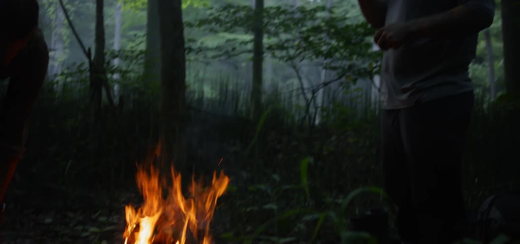 Man drinking water close to a bonfire in the middle of the forest