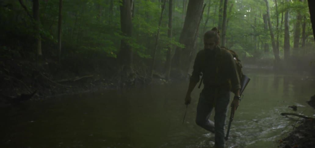 Man wades through shallow stream carrying rifle and backpack