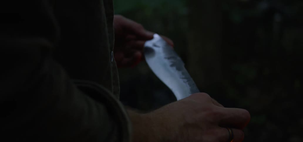 A man holds and inspects a metal blade while standing near a fire outdoors