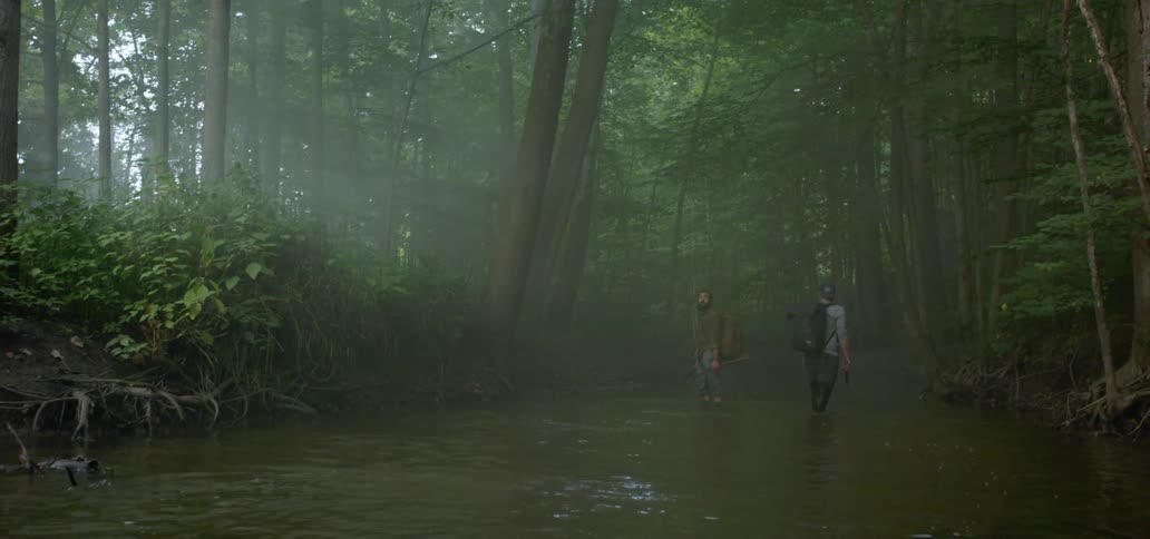 Men with backpacks crossing a shallow river while hiking on the woods
