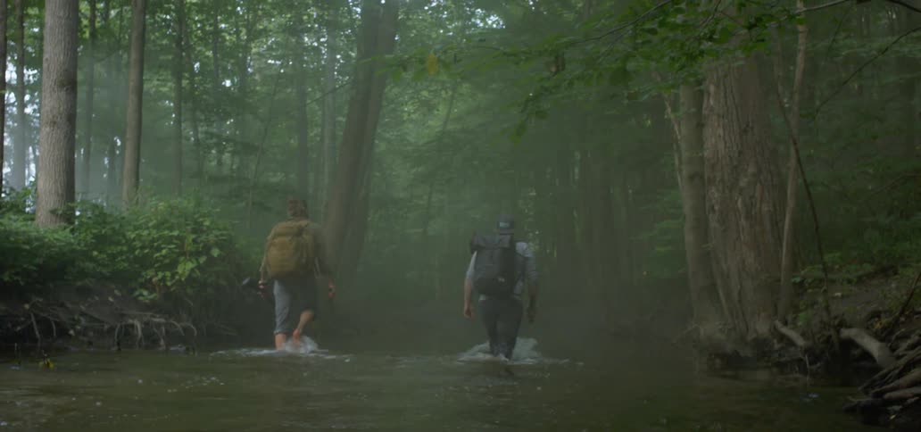 Two men with backpacks walk through knee deep waters of a stream flowing through a dense forest