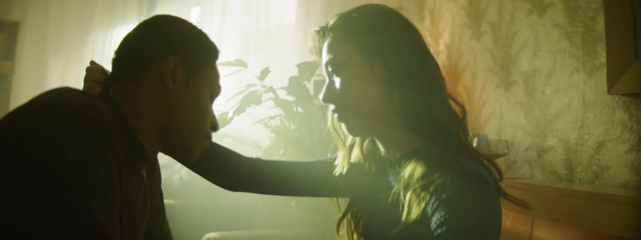 A young woman caresses the neck of a young man as they sit together indoors