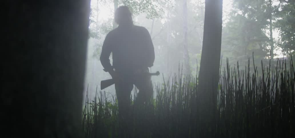 A man holds a rifle and walks through foggy woodland area