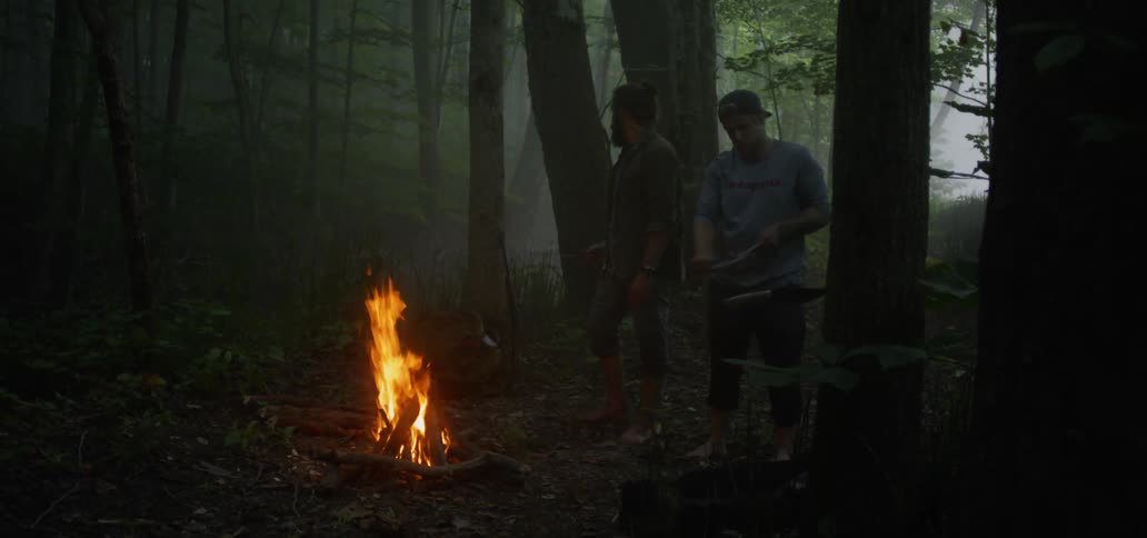 Two men standing over a campfire one whittling wood
