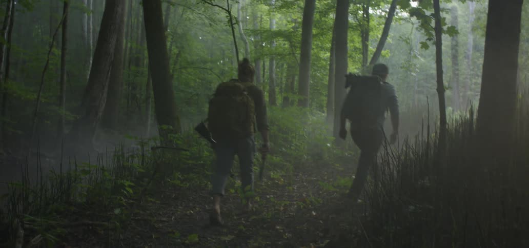Two men with backpacks walk through the woods while one carries a knife and a gun