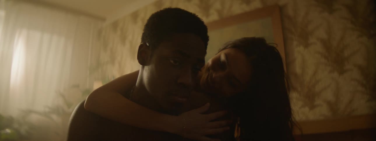 A woman hugs and kisses a man sitting on the edge of a bed