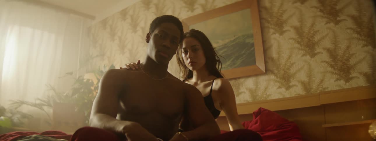Couple sitting shirtless on bed in bedroom looking at camera