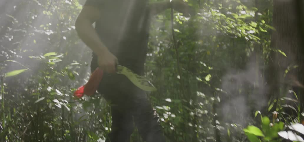 A man walks through a dense forest holding a small machete