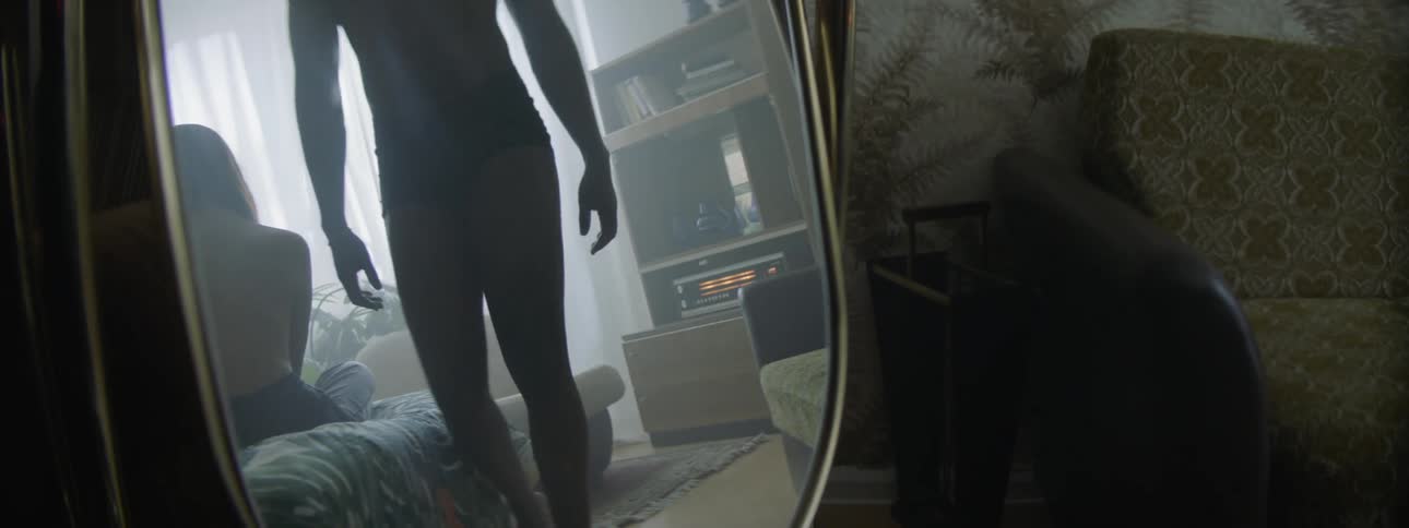 A female teen sits on a bed as a male teen in only his underwear gets up off a chair