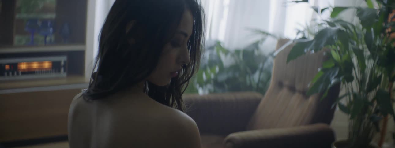 A long haired woman sitting in a room turns her head and looks down at her bare shoulder