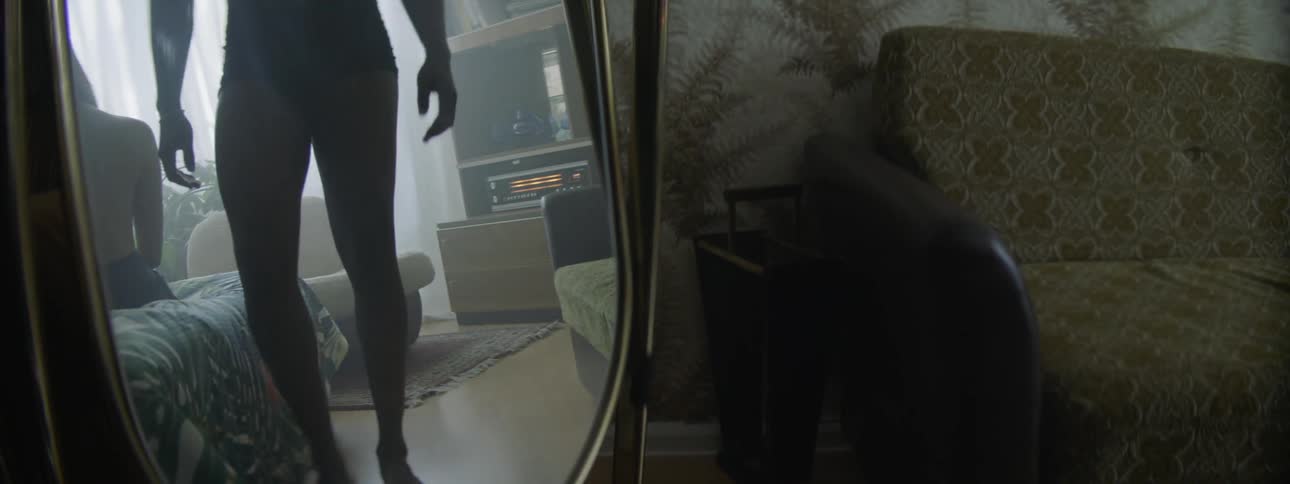 A man in his underwear rising from a chair and walking away as a woman sits on a bed
