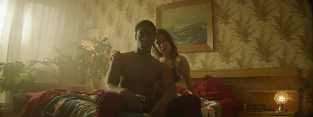 A young couple pose on a bed in a room with floral wallpaper
