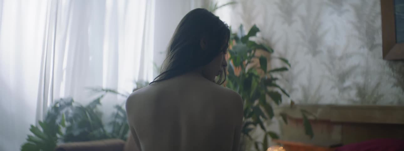 A woman sits topless at the end of a bed and turns her head