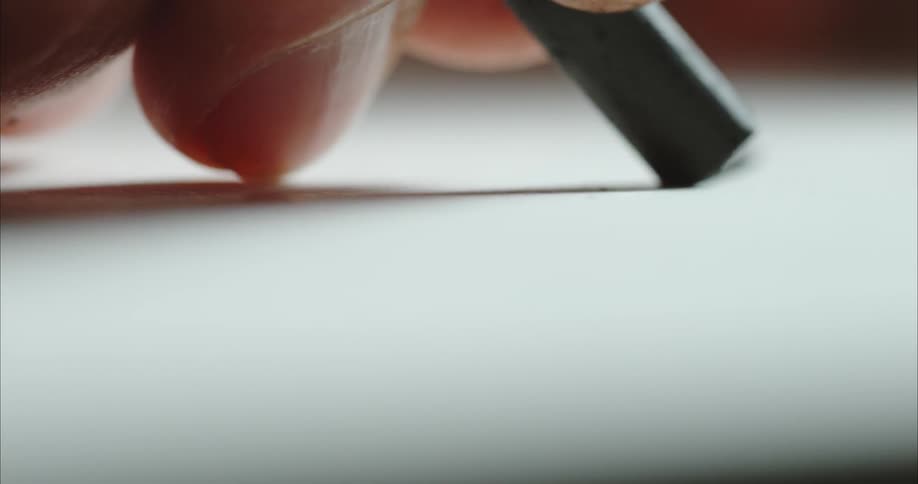 A person holding a stick of dark graphite uses it to draw a line across a piece of paper