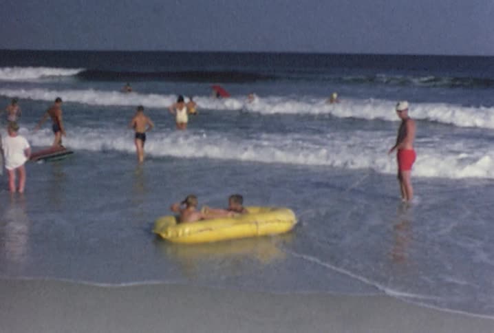 Old footage of people playing at the beach as a man holds a rope attached to a raft that two boys are sitting in