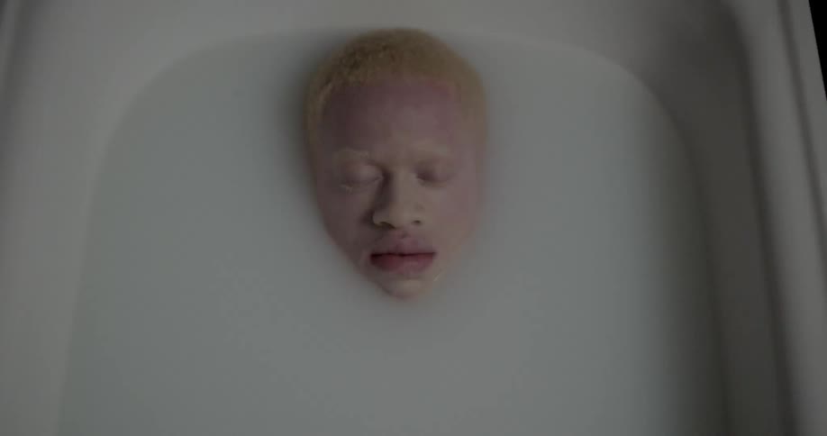 An young albino african male reclines in a bathtub of opaque milky water with only his face visible