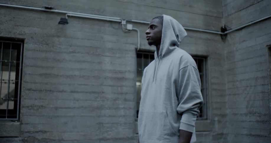 A man wearing a hoodie stands outside of a grey stone building