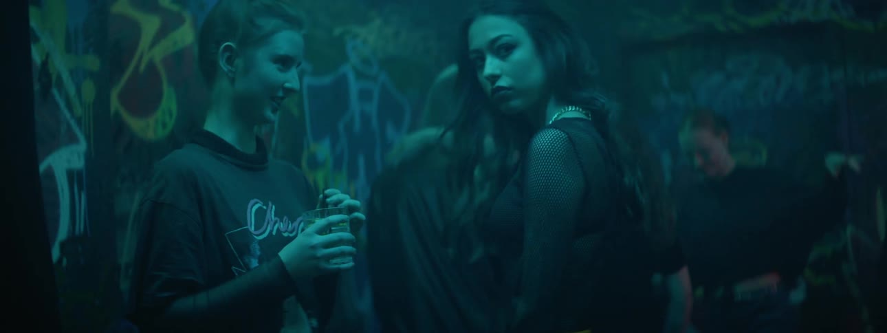 Two young women socialize within a graffiti covered dance club with strobing lights
