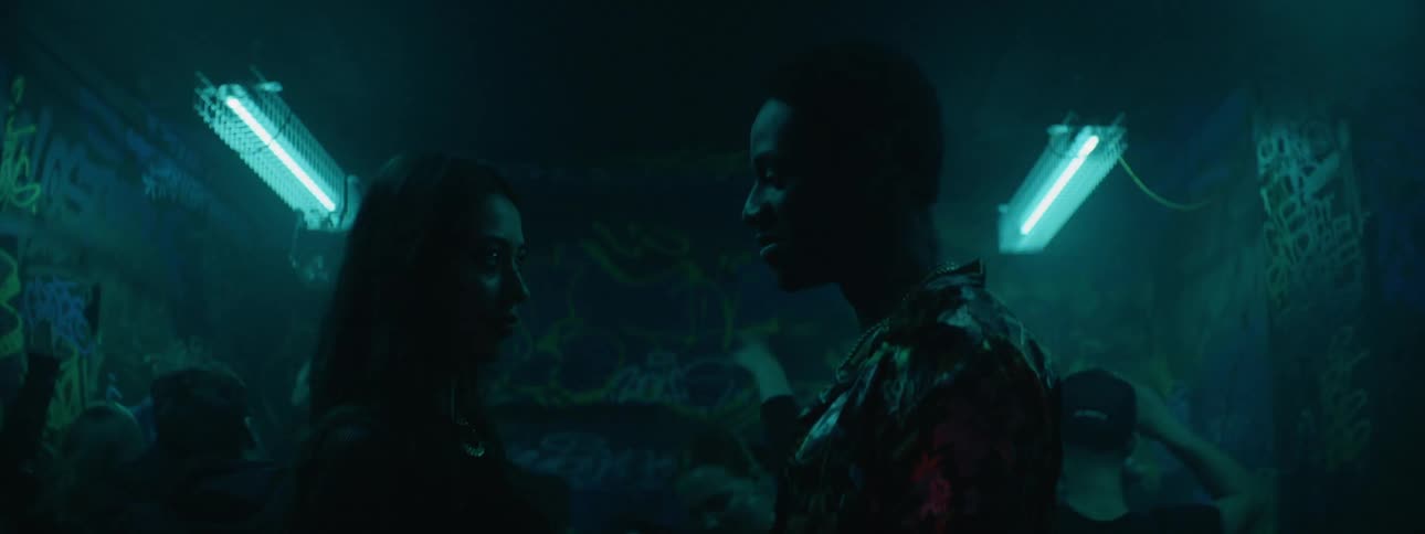 A young man and young woman stand facing each other in a dance club lit by strobing lights