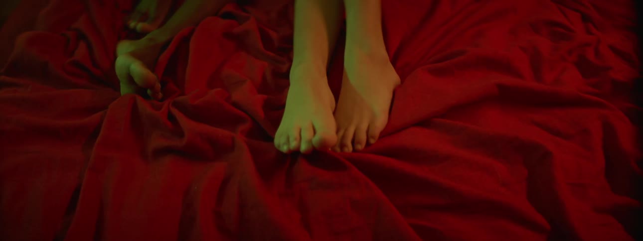 The bare feet of two people are seen rested atop a red blanket on a bed