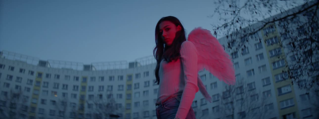 A young woman wearing furry pink costume wings stands before a large residental building
