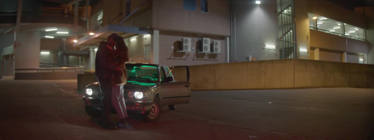 A man and woman kiss standing in front of a parked car at night
