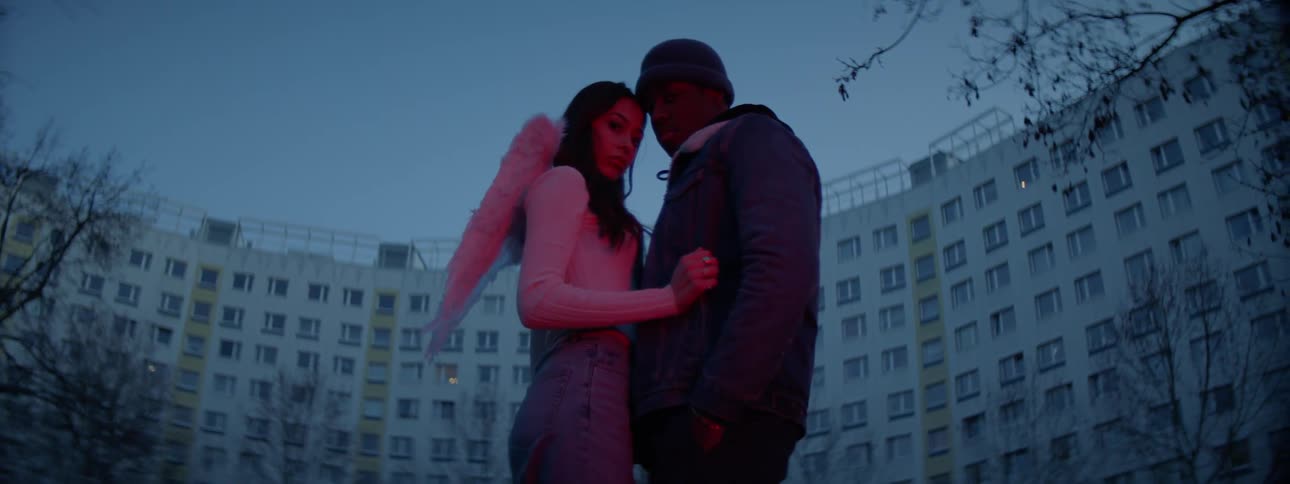 A woman with small angel wings on her back embracing a man in front of a large building in the evening